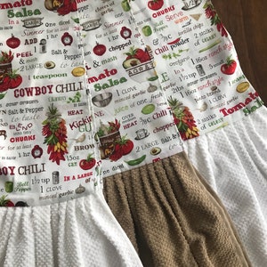 May include: Two white kitchen towels with brown trim. The towels have a red, green, and white print with text that reads "Cowboy Chili", "Tomato Salsa", and "Kickin' Guacamole".