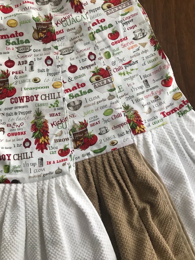 May include: Two white kitchen towels with brown trim. The towels have a red, green, and white print with text that reads "Cowboy Chili", "Tomato Salsa", and "Kickin' Guacamole".