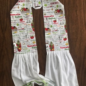 May include: White kitchen towel with green stripes. The towel features a printed design of a recipe for Cowboy Chili and Tomato Salsa. The recipe includes ingredients such as chili powder, cayenne pepper, and coriander.