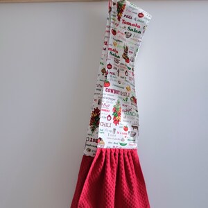 May include: Red kitchen towel with a white print of various food items and recipes. The print includes the words "Cowboy Chili", "Tomato Salsa", and "Kickin' Guacamole". The towel is hanging on a metal hook with the word "EAT" above it.