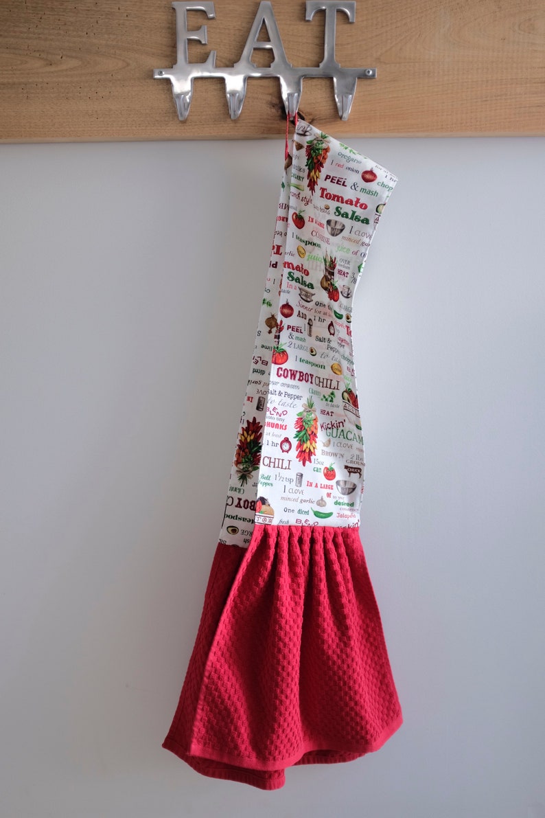May include: Red kitchen towel with a white print of various food items and recipes. The print includes the words "Cowboy Chili", "Tomato Salsa", and "Kickin' Guacamole". The towel is hanging on a metal hook with the word "EAT" above it.