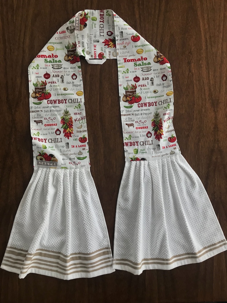 May include: Two white kitchen towels with brown stripes. The towels are printed with a red and green recipe for Cowboy Chili. The recipe includes ingredients like brown salt and pepper, ground beef, and chilli peppers.