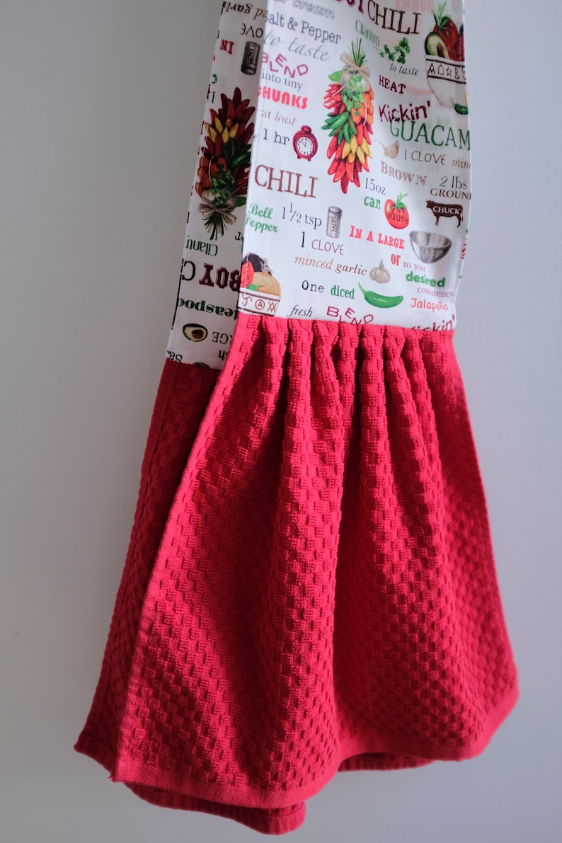 May include: Red kitchen towel with a white printed design featuring chili peppers, avocados, and text about chili and guacamole recipes. The text includes measurements in ounces and pounds.
