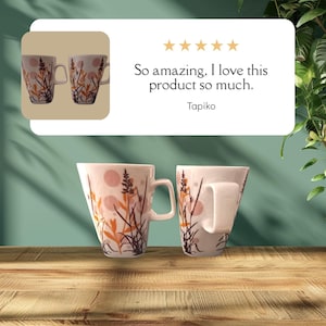 Puede incluir: Dos tazas de cerámica blanca con un diseño floral en naranja, marrón y negro. Las tazas tienen una forma única y un asa curvada. El texto "So amazing, I love this product so much." se muestra encima del nombre "Tapiko".