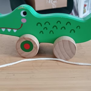 Könnte beinhalten: Ein grünes Holzspielzeug in Form eines Alligators zum Ziehen, mit einem lächelnden Gesicht, weißen Zähnen und Holzrädern. Der Alligator hat eine weiße Schnur. Ein lustiges Spielzeug für Kleinkinder.