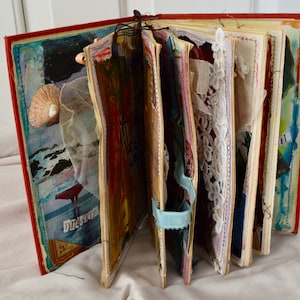 Op de afbeelding: Een rood, met stof bekleed dagboek met pagina's gevuld met mixed media kunst, waaronder verf, stofresten en kant. Het dagboek is open om de kleurrijke en getextureerde pagina's te laten zien.