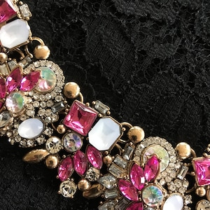 Pode incluir: Um colar de declaração em tom dourado com strass rosa e transparentes. O colar apresenta uma variedade de formas e tamanhos de pedras, incluindo quadrados, ovais e redondos.