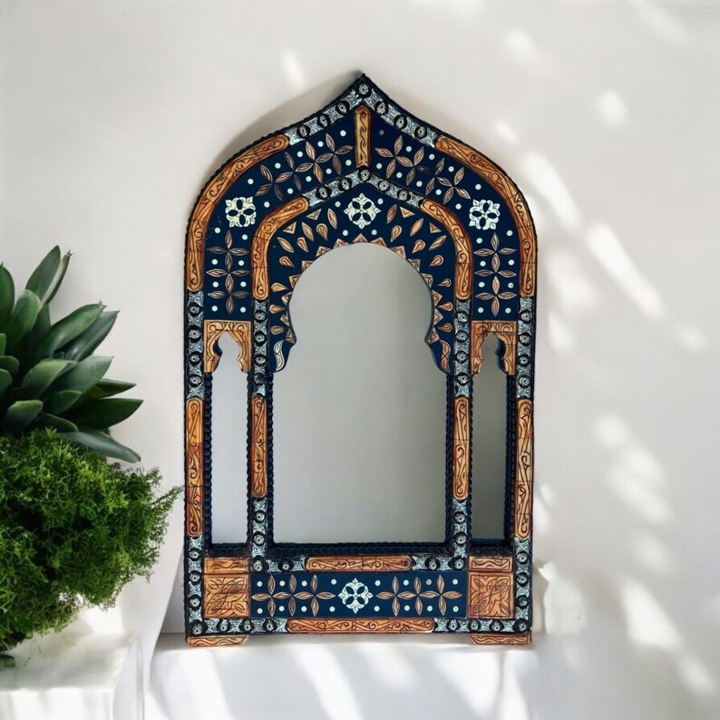 May include: An arched wall mirror featuring a dark blue frame with intricate light brown wood carvings and white floral patterns. The frame has a pointed top and side cutouts, creating a multi-tiered architectural silhouette with detailed geometric motifs.