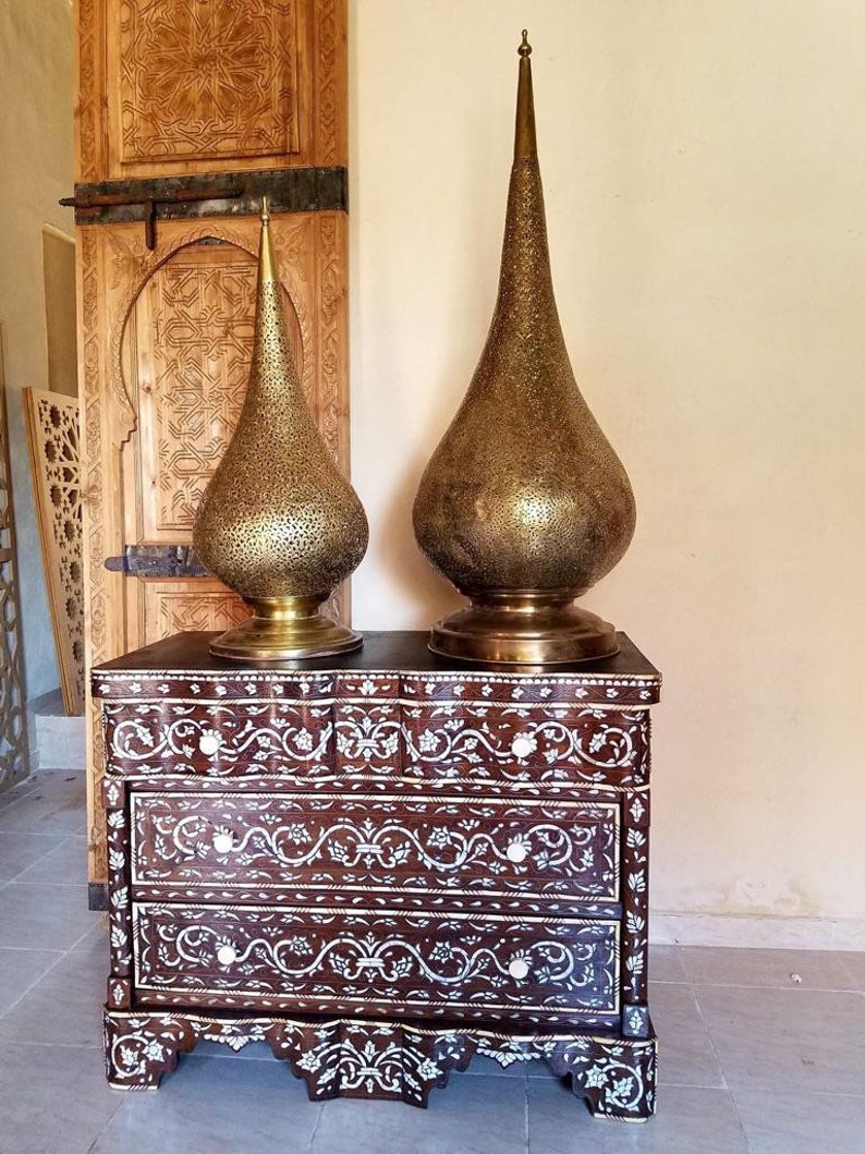 May include: Two ornate gold metal lamps with a teardrop shape sit on top of a dark brown wooden chest of drawers with three drawers. The chest of drawers is decorated with intricate floral patterns made of mother of pearl.