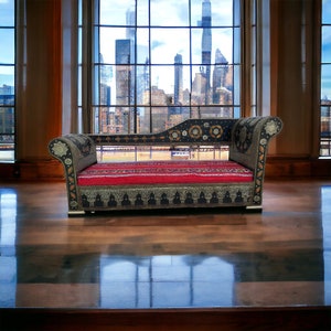 May include: A black and silver ornate couch with a red and white patterned cushion. The couch is in front of a large window with a view of a city skyline.