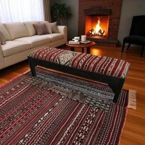 May include: A dark wooden bench with a patterned cushion in shades of red, brown, and cream sits atop a matching area rug. The rug and bench feature geometric designs. The scene is set in a living room with a fireplace.