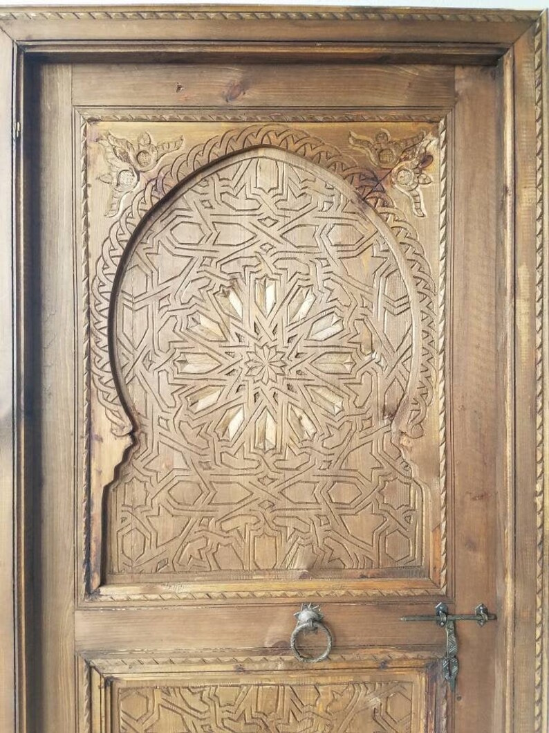 May include: A wooden door with intricate carvings in a geometric pattern. The door has a metal ring handle and a latch.