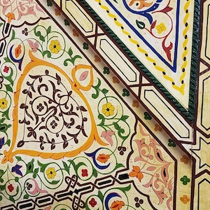 K&ouml;nnte beinhalten: Eine Nahaufnahme einer dekorativen Holzplatte mit aufwendigen handgemalten Designs. Die Platte zeigt ein geometrisches Muster mit einem zentralen Dreieck und floralen Motiven in den Farben Gelb, Orange, Rot, Blau, Gr&uuml;n und Braun.