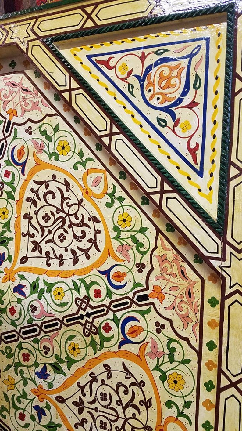 K&ouml;nnte beinhalten: Eine Nahaufnahme einer dekorativen Holzplatte mit aufwendigen handgemalten Designs. Die Platte zeigt ein geometrisches Muster mit einem zentralen Dreieck und floralen Motiven in den Farben Gelb, Orange, Rot, Blau, Gr&uuml;n und Braun.