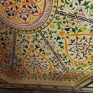 K&ouml;nnte beinhalten: Eine wei&szlig;e Decke mit komplizierten floralen Mustern, die in Gr&uuml;n-, Gelb-, Orange- und Blaut&ouml;nen bemalt sind. Das Design zeigt sich in sich wiederholenden Motiven von Bl&auml;ttern, Blumen und geometrischen Formen.