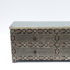 May include: A black and silver dresser with three drawers. The dresser has a glass top and is decorated with intricate silver patterns and red accents. The drawer pulls are dark metal.
