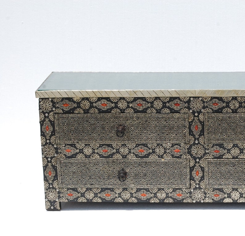 May include: A black and silver dresser with three drawers. The dresser has a glass top and is decorated with intricate silver patterns and red accents. The drawer pulls are dark metal.