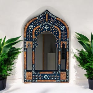 May include: An arched wall mirror featuring a dark blue frame with intricate light blue and tan floral patterns. The frame has a pointed top and decorative cutouts, with a central reflective glass panel surrounded by detailed, symmetrical carvings.