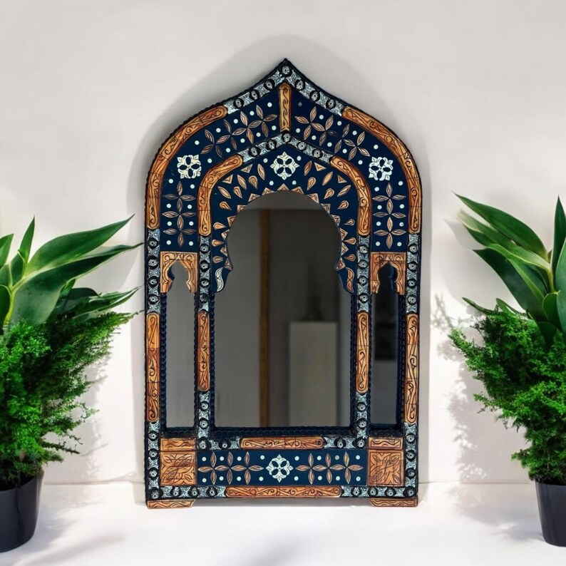 May include: An arched wall mirror featuring a dark blue frame with intricate light blue and tan floral patterns. The frame has a pointed top and decorative cutouts, with a central reflective glass panel surrounded by detailed, symmetrical carvings.
