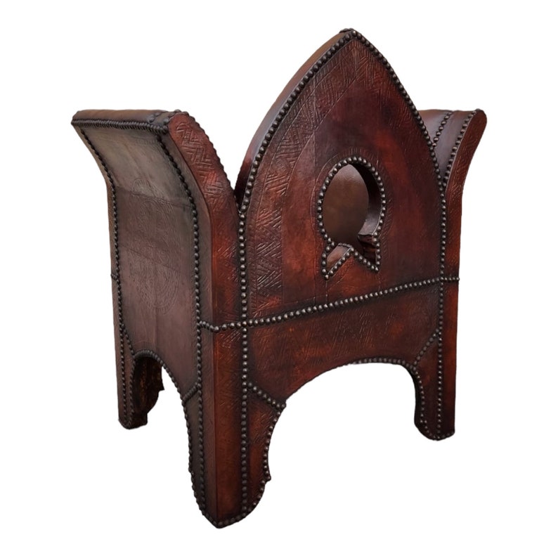 May include: A brown leather Moroccan pouf with a decorative cutout design and studded details. The pouf has a square base and a pointed top.