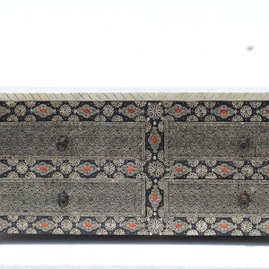 May include: A decorative chest of drawers with six drawers. The chest features intricate silver filigree designs on a black background, accented with orange and white floral patterns. The top is a light color with a textured surface.