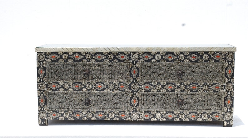 May include: A decorative chest of drawers with six drawers. The chest features intricate silver filigree designs on a black background, accented with orange and white floral patterns. The top is a light color with a textured surface.