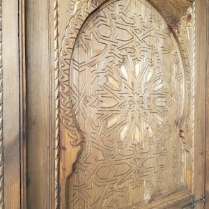 May include: A wooden door with intricate carvings in a geometric pattern. The door is painted a light brown color and has a metal handle.