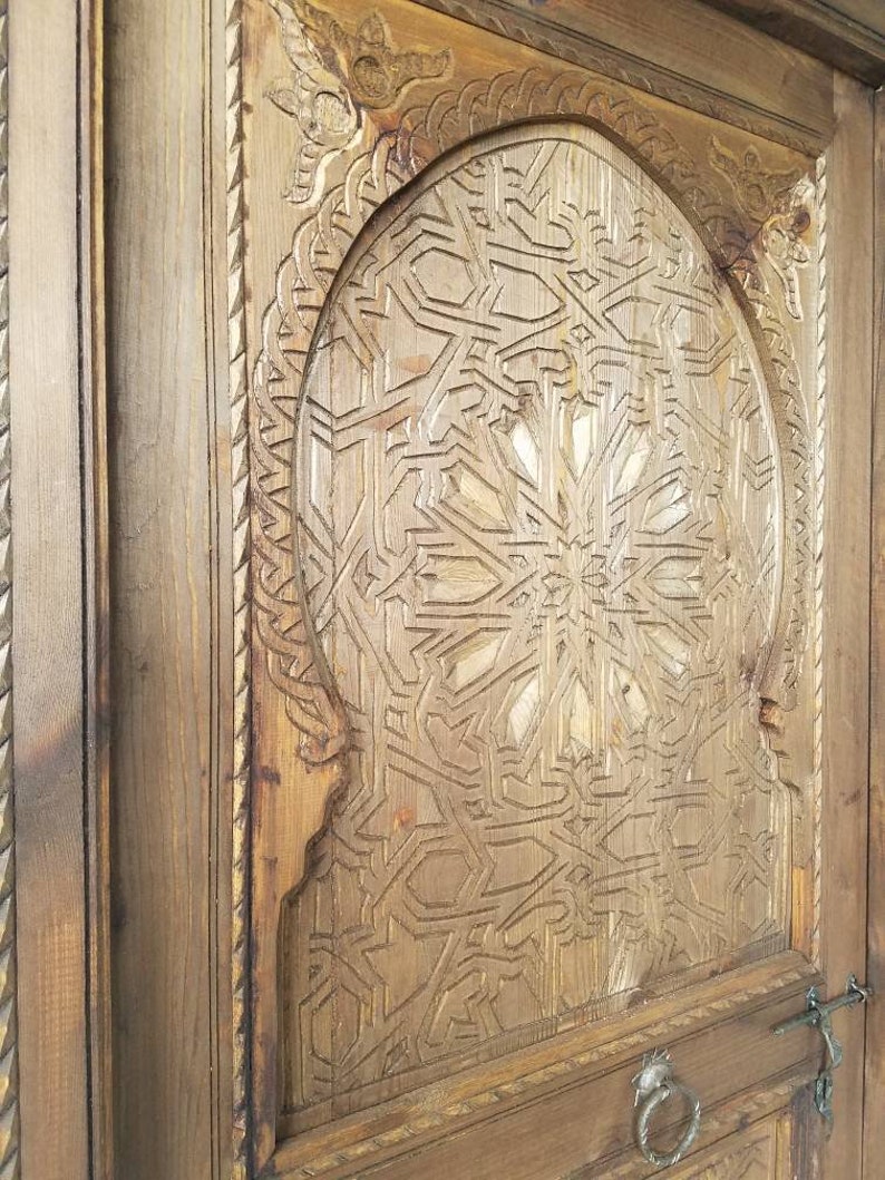 May include: A wooden door with intricate carvings in a geometric pattern. The door is painted a light brown color and has a metal handle.