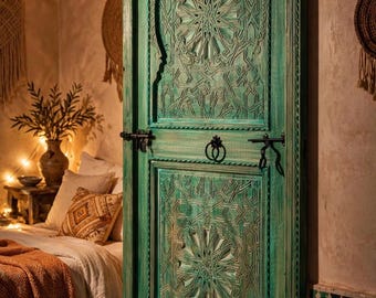 May include: A tall, rectangular wooden door finished in a distressed teal green paint. The door features intricate geometric carvings in two panels, a central metal ring handle, and two horizontal black metal sliding bolt latches.