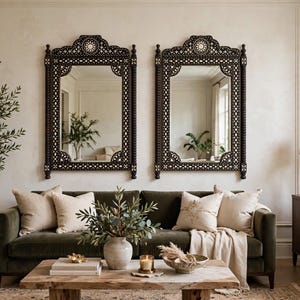 May include: Two rectangular wall mirrors with dark brown, intricately carved wooden frames featuring geometric patterns and white inlay details. The mirrors are positioned side-by-side above a dark green velvet sofa in a living room setting.