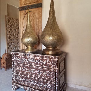 May include: A wooden chest of drawers with intricate floral inlays made of mother of pearl. Two tall, gold, decorative lamps with a teardrop shape sit on top of the chest.