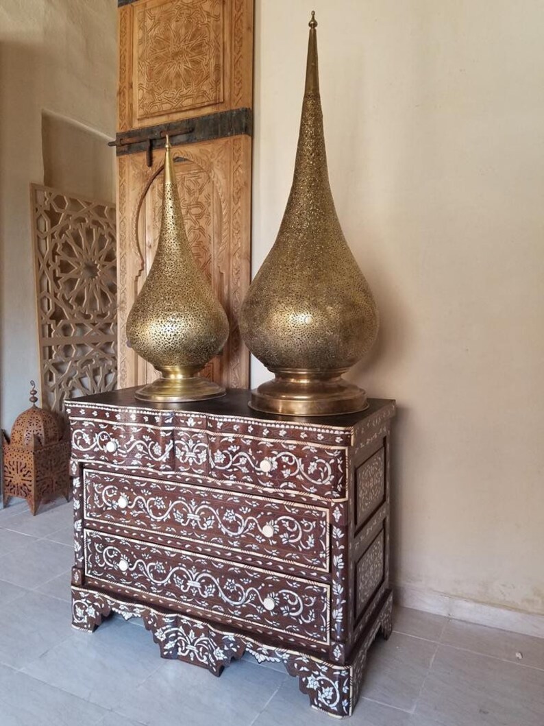 May include: A wooden chest of drawers with intricate floral inlays made of mother of pearl. Two tall, gold, decorative lamps with a teardrop shape sit on top of the chest.