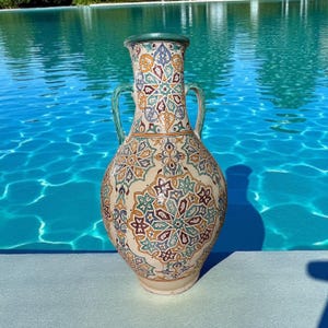 May include: A tall, decorative ceramic vase with a cream-colored base and intricate, colorful floral patterns in shades of blue, orange, and green. The vase has two handles and a green rim, standing against a backdrop of a blue pool.