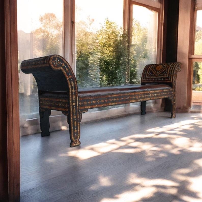 May include: A dark wood bench with intricate inlaid designs in gold and white. The bench has a curved back and is upholstered in brown leather. The bench is sitting in front of a window with a view of trees.