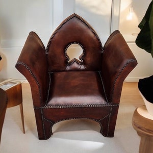 May include: A brown leather armchair with a distinctive, pointed back and a cut-out design in the back. The chair has a studded trim and a slightly distressed look.