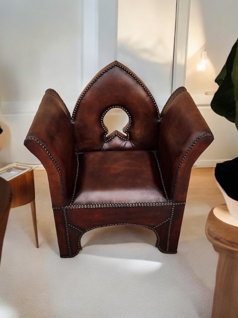 May include: A brown leather armchair with a distinctive, pointed back and a cut-out design in the back. The chair has a studded trim and a slightly distressed look.