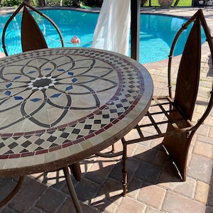 May include: A round, mosaic-tiled table with a geometric design in shades of brown, black, and white. The table is made of metal and has a rustic, weathered look. Two matching metal chairs with a similar design are placed around the table. The chairs have a curved back and a simple, openwork design.