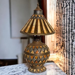 May include: An ornate table lamp with a decorative design. The lamp features a bronze base and shade with cream and brown accents. The base has a spherical shape with diamond and triangle patterns. The shade has a conical shape with vertical stripes.