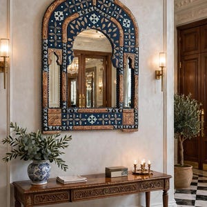 May include: An arched wall mirror featuring a dark blue frame with intricate light-colored floral patterns and carved wood accents. The mirror has a pointed top and a rectangular base, with a reflective glass surface that shows a room interior.