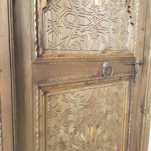 May include: A close-up of a wooden door with intricate carved designs. The door is made of light brown wood and has a dark brown stain. The carvings are in a geometric pattern and are very detailed. The door has a metal handle and a metal latch.