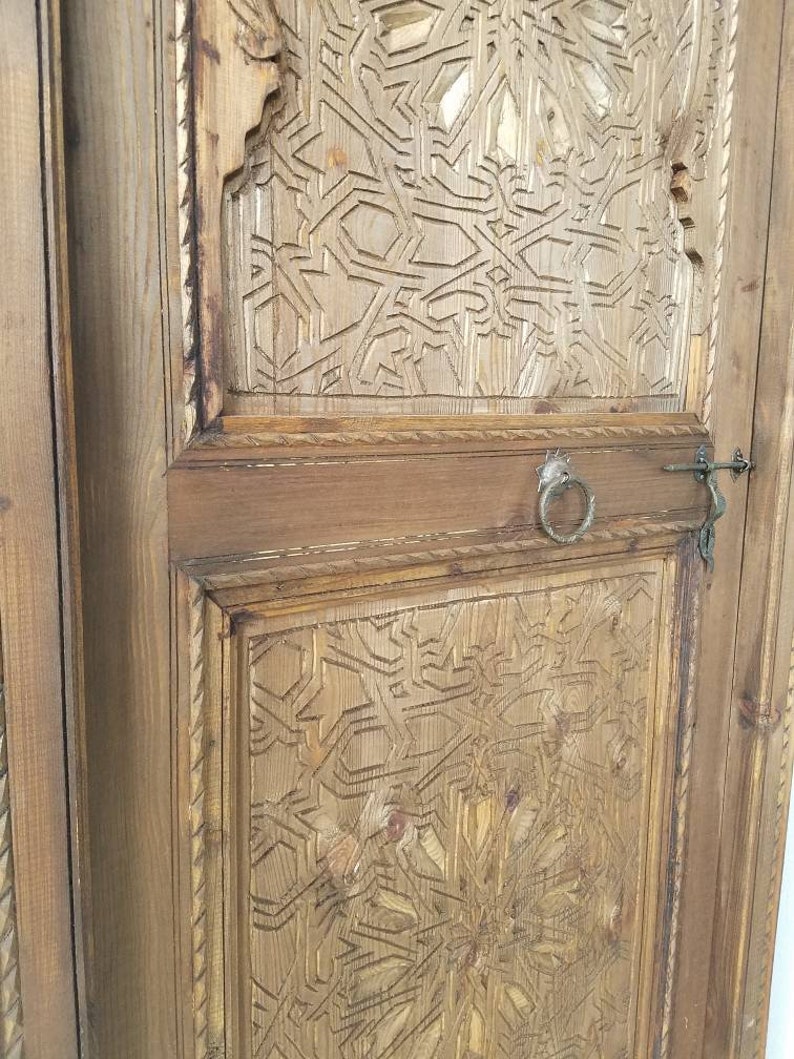 May include: A close-up of a wooden door with intricate carved designs. The door is made of light brown wood and has a dark brown stain. The carvings are in a geometric pattern and are very detailed. The door has a metal handle and a metal latch.