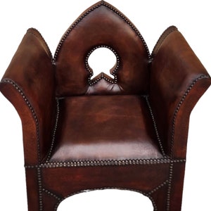 May include: A brown leather armchair with a distinctive, pointed back and a decorative cutout in the centre. The chair is studded with metal accents and has a traditional design.