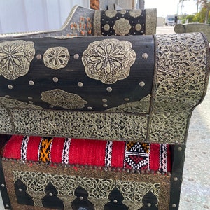 May include: A black wooden sofa with intricate silver metalwork and a red and white patterned cushion. The sofa has a curved back and armrests and is decorated with floral and geometric designs.