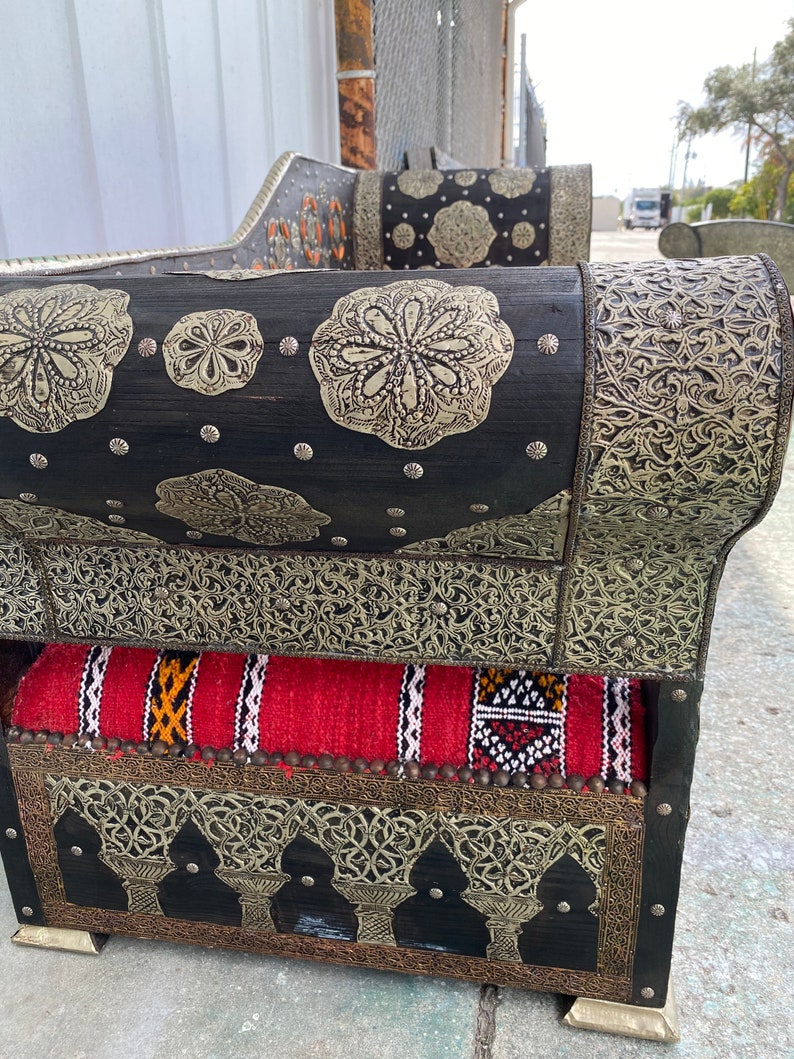 May include: A black wooden sofa with intricate silver metalwork and a red and white patterned cushion. The sofa has a curved back and armrests and is decorated with floral and geometric designs.