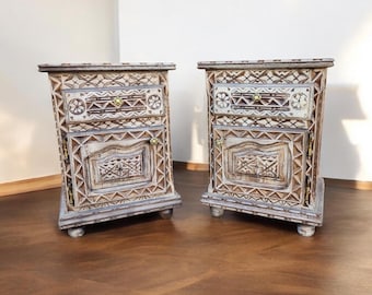 May include: Two white painted wooden nightstands with intricate carvings and a single drawer. The nightstands have a distressed finish and are sitting on a wooden floor.
