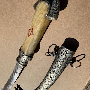 May include: A silver and bone dagger with an ornate handle and a scabbard with intricate floral designs. The dagger has a red and white emblem on the handle.