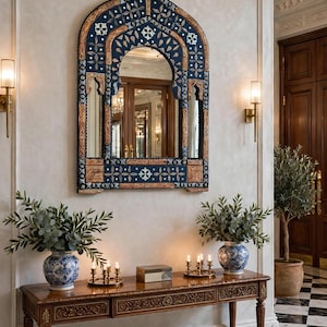May include: An arched wall mirror with a dark blue frame featuring intricate gold and white floral patterns, mounted above a rectangular wooden console table with carved details, two blue and white ceramic vases, and brass candle holders.