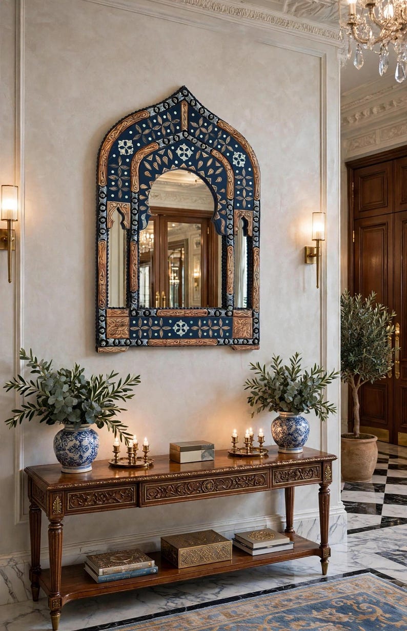 May include: An arched wall mirror with a dark blue frame featuring intricate gold and white floral patterns, mounted above a rectangular wooden console table with carved details, two blue and white ceramic vases, and brass candle holders.