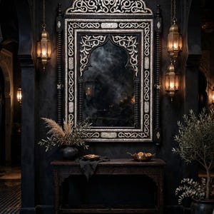 May include: A rectangular wall mirror featuring an intricate silver-toned frame with ornate scrollwork and a pointed arch top. The mirror is mounted above a dark wooden console table, flanked by two hanging brass lanterns with warm glowing lights.