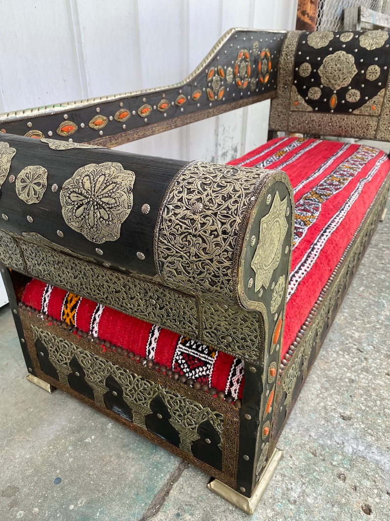 May include: A black wooden couch with intricate silver metalwork and red and white patterned cushions. The couch has a high back and rolled arms. The couch is decorated with small orange gemstones.